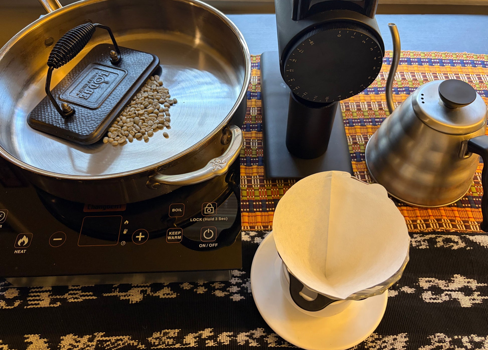 A coffee roasting and brewing setup on a patterned table runner, featuring a stainless steel pan with a cast iron press and raw beans, a black electric grinder, a stainless steel gooseneck kettle, and a white paper dripper on a saucer.