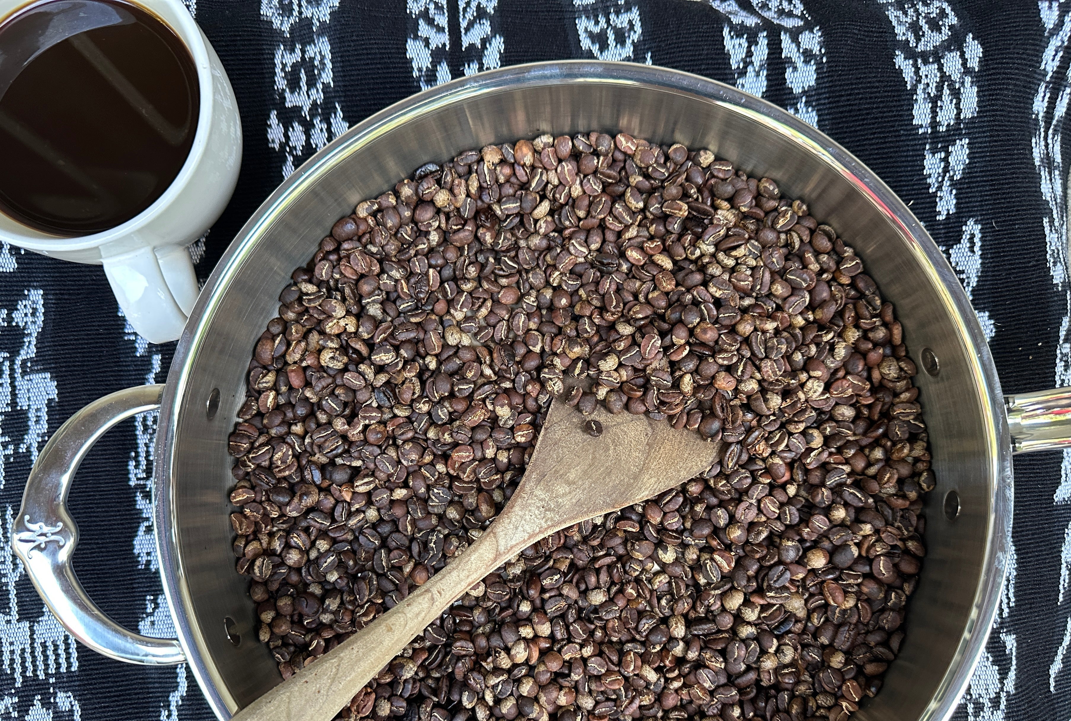 A top-down view of roasted coffee beans in a pan with a wooden spoon, next to a white mug of coffee on a black and white patterned textile.